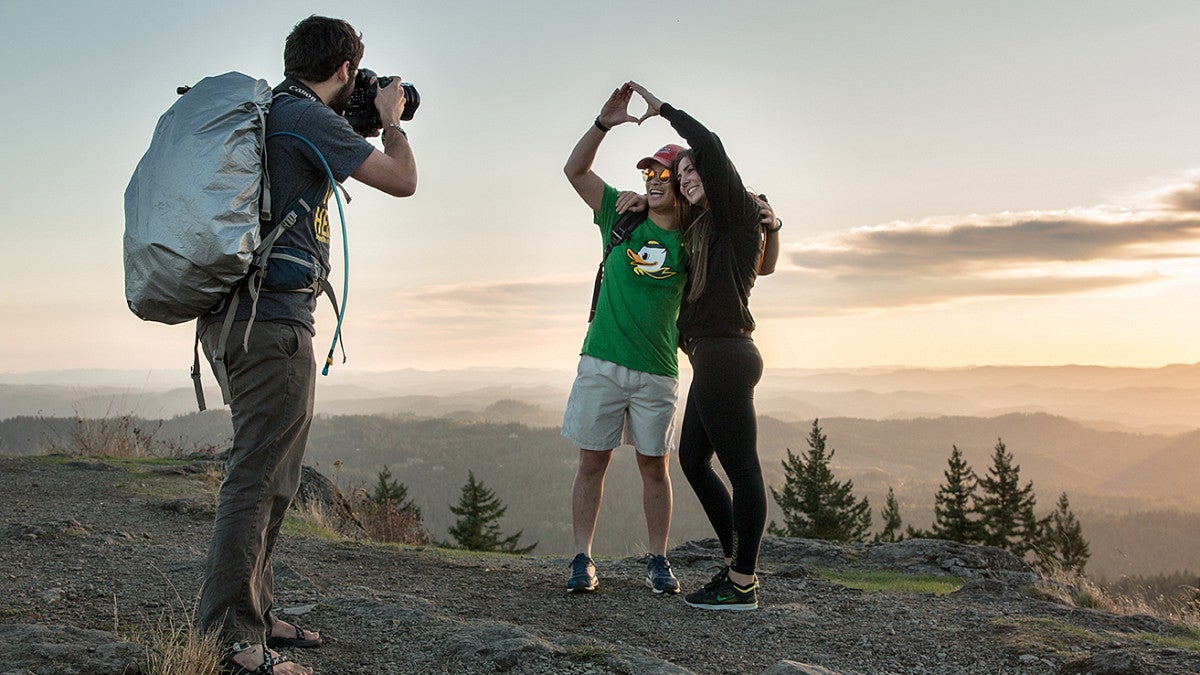 Sunset photo of one person taking a photo of two people on a hill "throwing the 'O'" during a photo session 